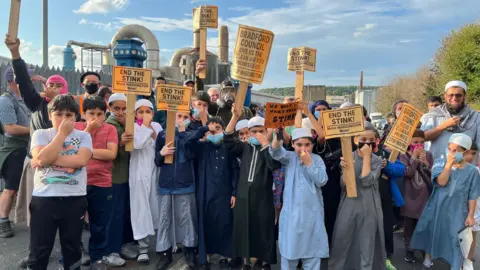 Aisha Iqbal/BBC A large crowd of children, all boys, with a few adults peppered within the group. Most of the children hold their noses and have a disgusted look on their face. Some of them wear face masks. Others hold home made placards saying things like 'end the stink' and 'give us the clean air you promised'.
