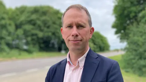 Matthew Barber has short brown hair, and is wearing a pink shirt and blue blazer. There is a road out of focus behind him.