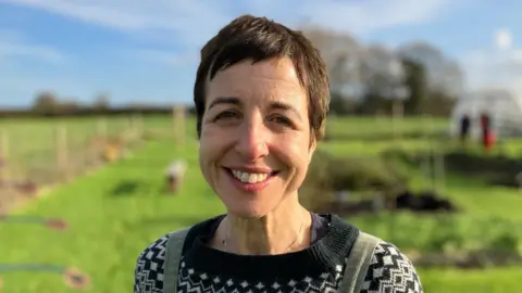 Dr Susan Taheri - a woman in dungarees with black and white jumper in a surgery garden with people behind her.