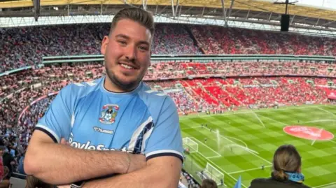 Matthew Loveitt smiling looking towards the camera wearing a sky blue Coventry City shirt at Wembley Arena.