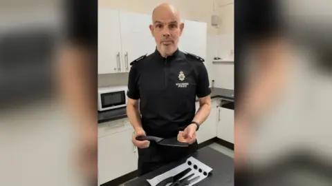Wiltshire Police A man with balding hair and a grey beard wearing a black Wiltshire Police uniform holding a rounded-tip kitchen knife above a table with a packet of three more rounded-tip kitchen knives with a white kitchen in the background. 