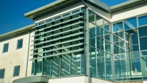 Blaenavon Medical Practice with large glass panels above its door. It is a modern-looking building with a cream exterior to the left and large glass panels to the right, with vertical beams and horizontal shutters making a glass grid in the middle. 