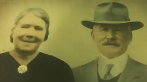 Campbell family Malcom is pictured with a woman. He has a moustache and is wearing a fedora, a shirt and tie and a suit.