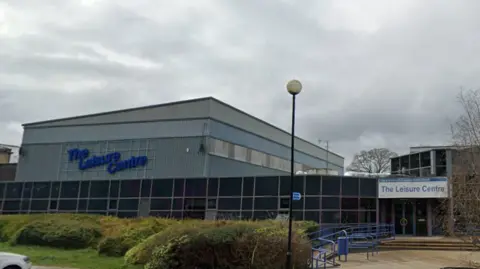 The front of the leisure centre, with ramps leading up to the entrance on the right, and a large blue sign on the hall to the left.