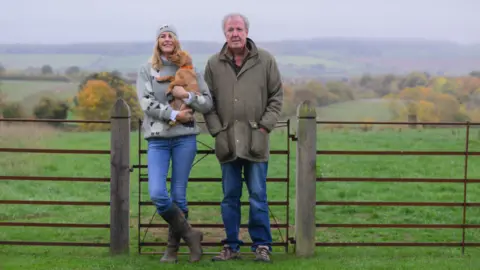 Farm-Fest A woman with long blonde/brown hair and wearing a grey jumper is next to a man in a coat. They are both wearing blue jeans and standing by a gate in a field.
