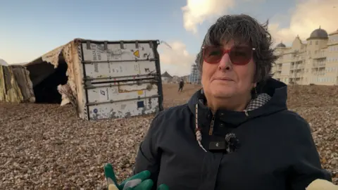 A woman with short black hair and red sunglasses stood on a pebble beach looking to the left of the camera. Behind her is a large white shipping container which is on the beach and has been badly damaged