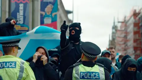 Masked protesters and police officers face each other in a city street during a public demonstration. Several people wear dark clothing and face coverings, while two police officers in high-visibility jackets stand in the foreground. A large blue-and-white vehicle or display is visible behind the crowd, with city buildings and scaffolding in the background.