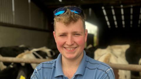 Ryan Taggart standing with his arms crossed looking at the camera. In the background there are dairy cows eating in a shed.