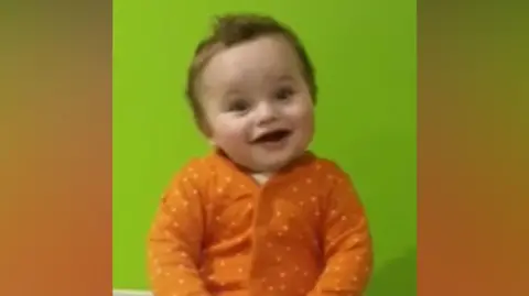 Baby in orange and white polka dot baby grow smiling while sitting on a changing table. Green wall behind him. 