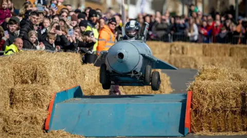 Borough Council of King's Lynn and West Norfolk A kart that looks like a jet jumping over a ramp with hay bales either side and dozens of spectators looking on