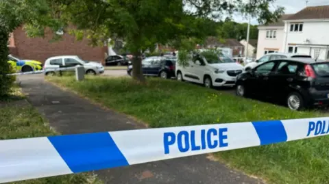 Blue and white police tape stretches across the image with cars and houses in the background. 
