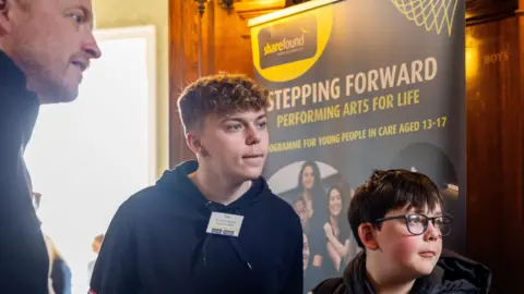 Ben Gregory-Ring/Tring Park School Three people looking off to the right, as though they are listening to someone speak. You can see a man with stubble, a teenage boy with short curly hair and Matthew. They are beside a sign that says Stepping Forward. 