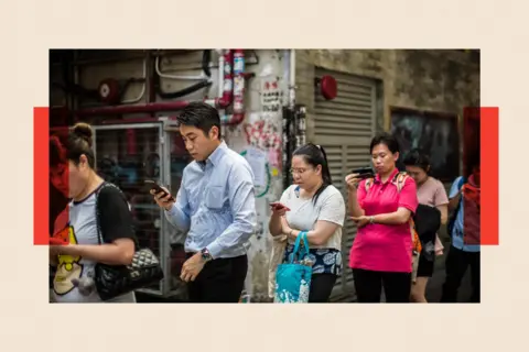 AFP via Getty Images Commuters look at their mobile phones as they wait in line for a bus after finishing work in Hong Kong 