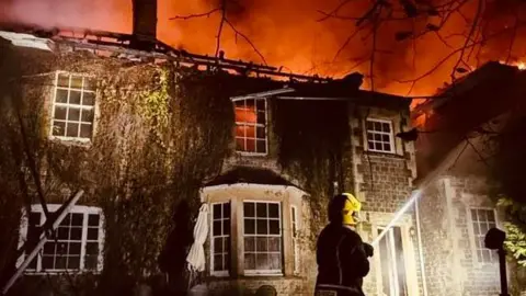 A georgian-style house at night on fire, the sky a reddish orange from the flames, the roof destroyed and the red can been seen through the windows. A firefighter is in front, using a hose.