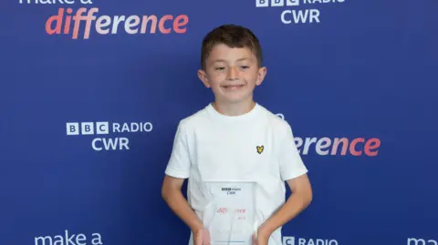 Mark Pemberton A young boy stands in front of a blue background holding a make a difference award