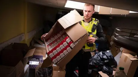 Lincolnshire Police A male police officer wearing black police uniform and a yellow hi-visibility vest holding a cardboard box that is filled with cigarette boxes. He is surrounded by boxes of cigarettes on the floor and a ceiling tile is loose above him.