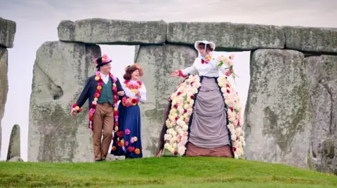 English Heritage A tall woman wearing a large skirt which is decorated with dahlias. She is wearing Victorian style clothing and a bonnet, and is walking alongside a couple with her arms outstretched and carrying a bouquet of dahlias. The couple are linking arms, also wearing Victorian clothing adorned with colourful dahlias.