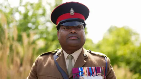 PA Media A man in green military uniform, wearing a significant number of medals on his chest. He has a black hat with a red band around its rim. There are trees in the distance behind him.