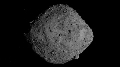 NASA/Goddard/University of Arizona gambar asteroid selebar 500m bernama Bennu yang terlihat seperti batu abu-abu yang lebih lebar di tengah daripada di setiap ujung. Tidak halus - ada gumpalan menonjol berukuran berbeda yang mencuat dari permukaannya. 