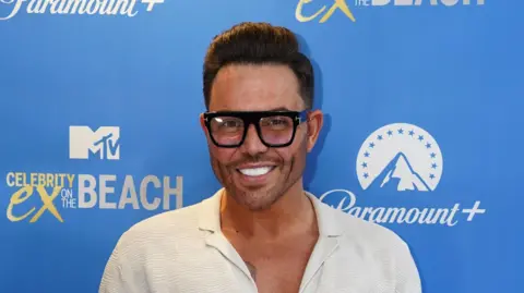 A man wearing a white shirt. He has thick frame black glasses and smiling at the camera.