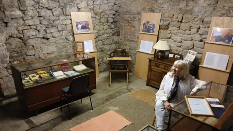 Supplied Elaine Humes siting next to a glass table in the stone-walled room of the future museum. She is surrounded by a glass cabinet and shelves with photographs. 