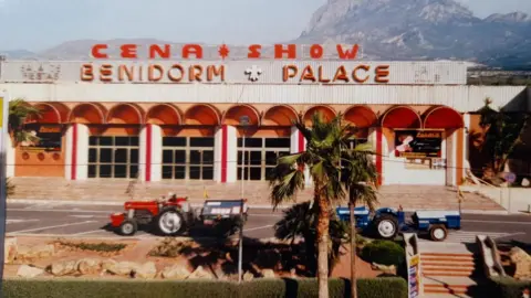 Dave Harrison Two tractors - one red and one blue - drive on a road past a large building with a sign reading : "Cena Show Benidorm Palace". There is a palm tree in the foreground and the weather is sunny.
