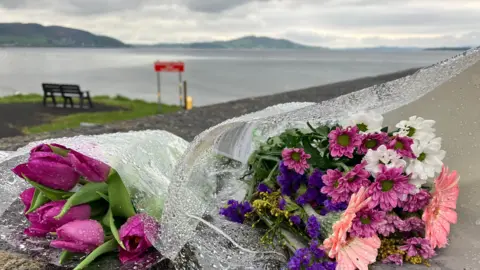 Pink, purple and white flowers wrapped in clear plastic are laid on the ground near the beach. In the background you can see a chair overlooking the sea with the hills in the background.