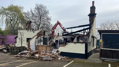 Tony Fisher/BBC A partially demolished single-storey building stands with much of its roof removed and interior exposed. A red mechanical excavator is positioned behind the structure, actively dismantling remaining sections of the roof and walls. Piles of broken bricks, timber, and debris are scattered across the ground in front of the building. The surviving walls show fire damage or heavy wear, with darkened, charred areas visible around window openings and roof beams. Nearby, a small outbuilding remains intact, and trees without dense leaves rise behind the site under an overcast sky.