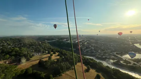 The Bristol landscape seen from a hot air balloon. The Clifton Suspension Bridge can be seen in the distance over Avon Gorge, as well the fields surrounding the city. Hot air balloons can be seen flying over the landscape, while the sun rises.