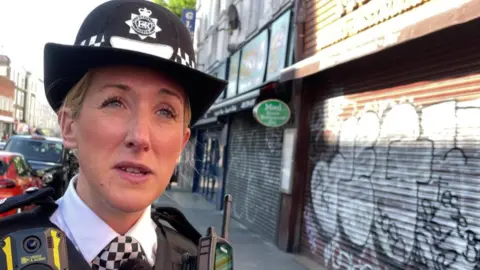 Brittany Clarke wears a police uniform, she has blonde hair and is standing in front of some shops with their shutters closed on a high street 