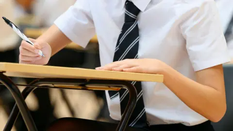 Student in school uniform writing on paper at a desk in a classroom, with other students blurred in the background.