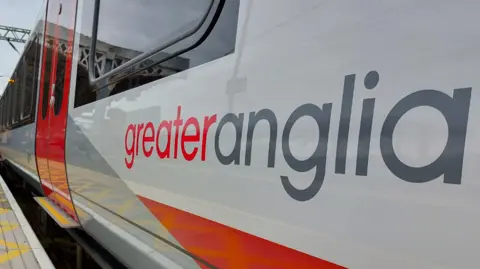 A silver Greater Anglia train that has the company's logo on the side in red and grey. The train is stopped at a platform.