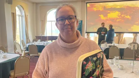 Emma Walton is wearing a pale pink polo-neck jumper and is smiling at the camera. She has her blonde hair tied back and is wearing glasses and holding a multi-coloured plastic folder. She is standing in a meeting room that has formal dining tables and chairs placed throughout. There is a large screen with a photo of an old type of aircraft flying over the island. Beneath the screen two men ate talking to each other.