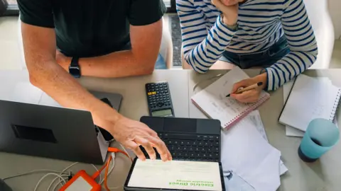 A couple sit at a large table in front of a laptop with a calculator and notebooks. They look to be going over a budgets and finances. The man is dressed in a dark t-shirt while the woman has a long-sleeved striped top.