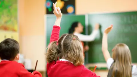 Girl with a ponytail in red school uniform. She has her hand up waiting to ask a question to the teacher at the front.
