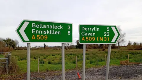 Green road signs for Enniskillen and Derylin. Picture taken outside during the day. Grey skies. Field in background. 