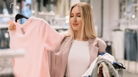 Getty Images A young woman with long straight brown hair in a clothes shop with a pile of tops on hangers over one arm and holding up a baby pink t shirt to look at in the other