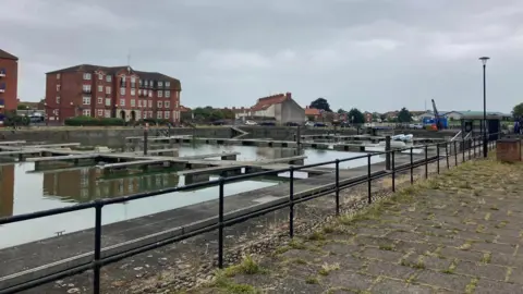 The docklands in Bridgwater. Wooden moorings can be seen in the water next to black railings on a concrete path. In the distance there are two large buildings.