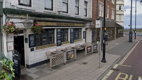 Google The Turks Head is a white stone terraced building with wooden garden furniture out front. Chalkboards are mounted on the front, detailing various offers. A gold and black sign above the windows reads: "Turks Head Hotel".