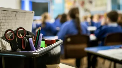 A generic image of a classroom. The backs of children who are sitting facing the front of the classroom are out of focus and a box of stationery in the foreground is in focus. 