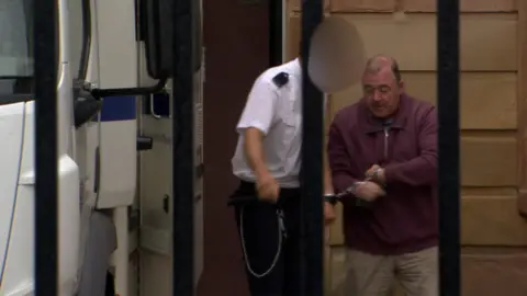 Fr Edward Gallagher being taken out of court in handcuffs. He is wearing a burgundy half-zip and beige trousers. He is accompanied by a prison officer.