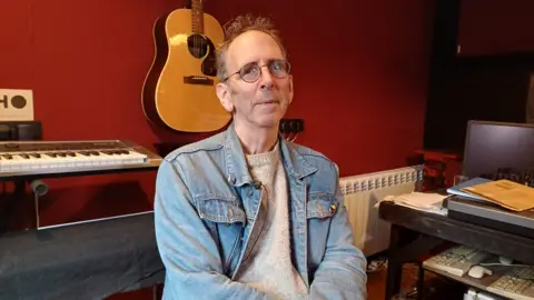 Gordon Maclean has fair hair and wearing glasses. He is wearing a denim jacket over a oatmeal-coloured jumper. He is sitting in a studio. Behind him are a keyboard and a guitar.