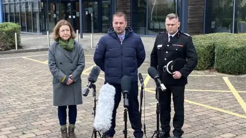 Chloe Turner, Leader of Stroud District Council, Det Supt Ian Fletcher and  deputy chief fire officer Nathaniel Hooton. They are standing in front of five microphones outside the police headquarters.
