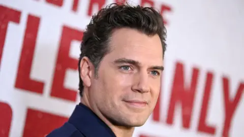 Getty Images A handsome man stands in front of a red carpet hoarding, smiling as he poses for pictures. The catchlight of camera flashes can be seen in his eyes.