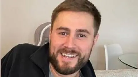 Mathew Hardy wearing a black and grey zip sweater. He is sitting somewhere smiling and has a beard with short, brown hair.