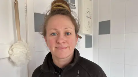 Woman with a ponytail on top of head smiles at camera with a bathroom behind