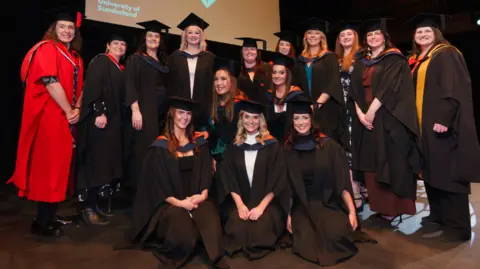 The first cohort of graduating midwives smile in their graduation black gowns and caps. They are all women of different ages. 
