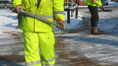 Two highways workers wearing high-vis clothing hold shovels with grit and sand on them as they spread it out across an icy road.