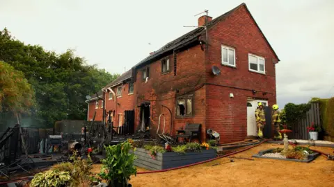 South Yorkshire Fire & Rescue Two firefighters stood at the door of a semi-detached property which has been badly fire damaged. The windows have shattered and the veranda in the garden is completely black from fire damage.