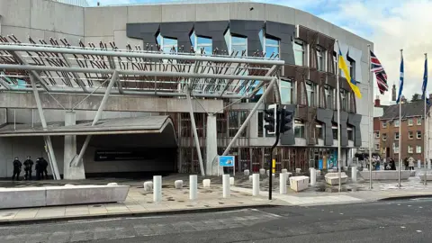 A modern concrete building - Holyrood- with irregular windows, wooden slats, a steel canopy at the entrance, bollards along the front, and several flags flying to the right.